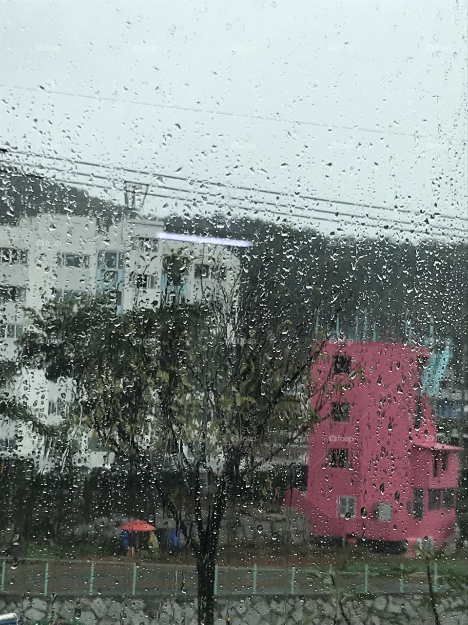 Outside you can see three people. They are under the pink big umbrella wearing an yellow and blue rain coat and doing some work for their little farm.