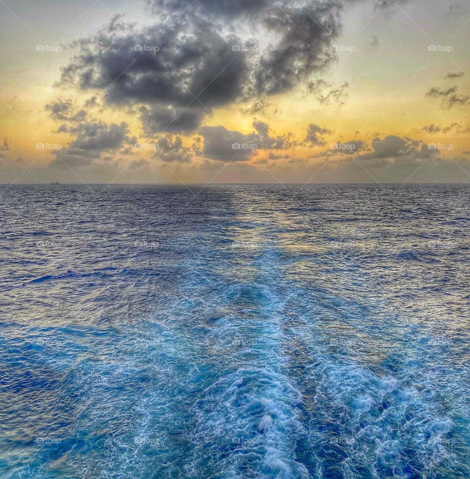 The view from our balcony at the back of a cruise ship.  It’s golden hour - aka sunset - and Mother Nature did not disappoint.  The horizon over the water is dotted with clouds, as the sky turns yellow and orange.   