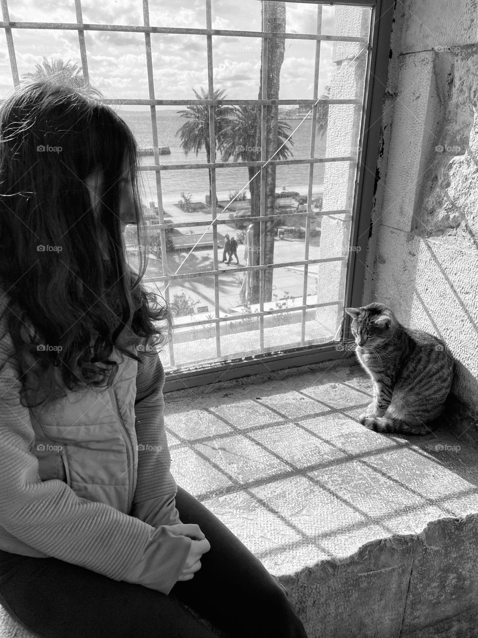 Black and white picture of a girl sitting in the window looking at a cat.