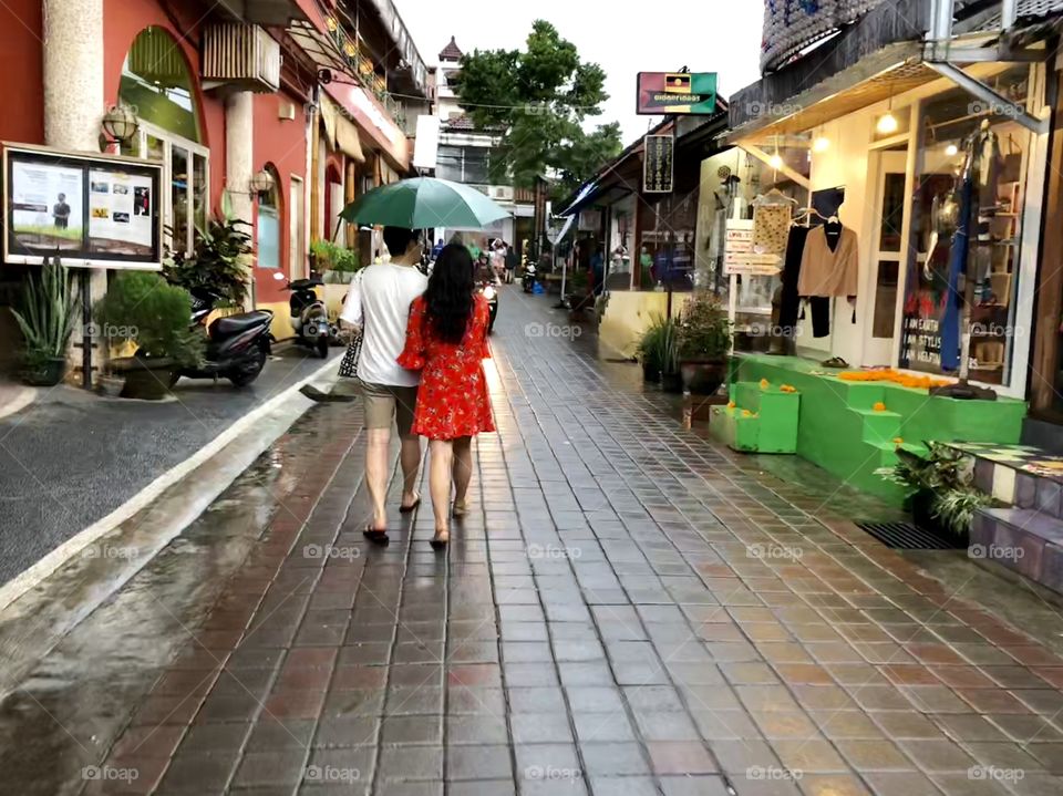 Walking in stride 3. Adult couple with open umbrella. Brick paved street. Shops. Ubud, BalI. 2018.