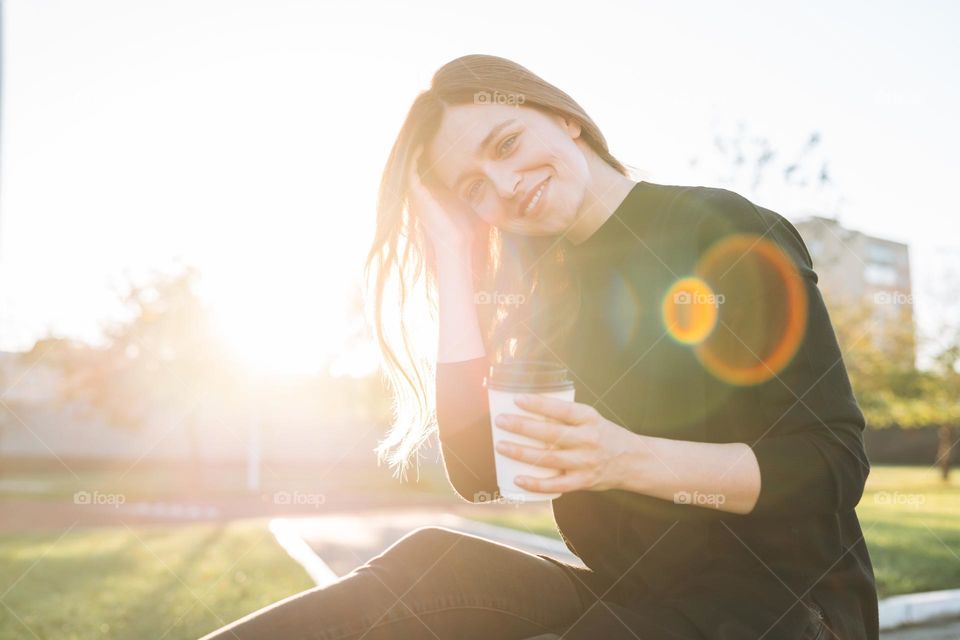 Portrait of young happy woman with long hair with paper cup of coffee in city park on golden hour