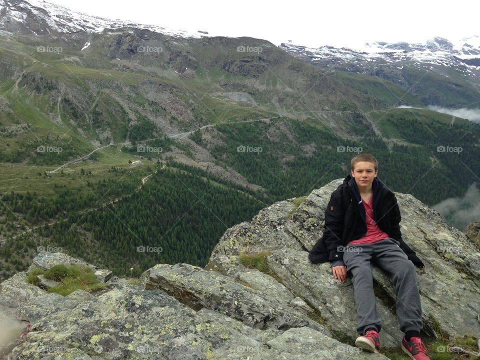 A teenage boy sitting on a rock by the edge of a shear cliff of a mountain in Zermatt, Switzerland. Filled with greenery in the background, it is roughly a mile away from the edge that the teenage boy is resting on.