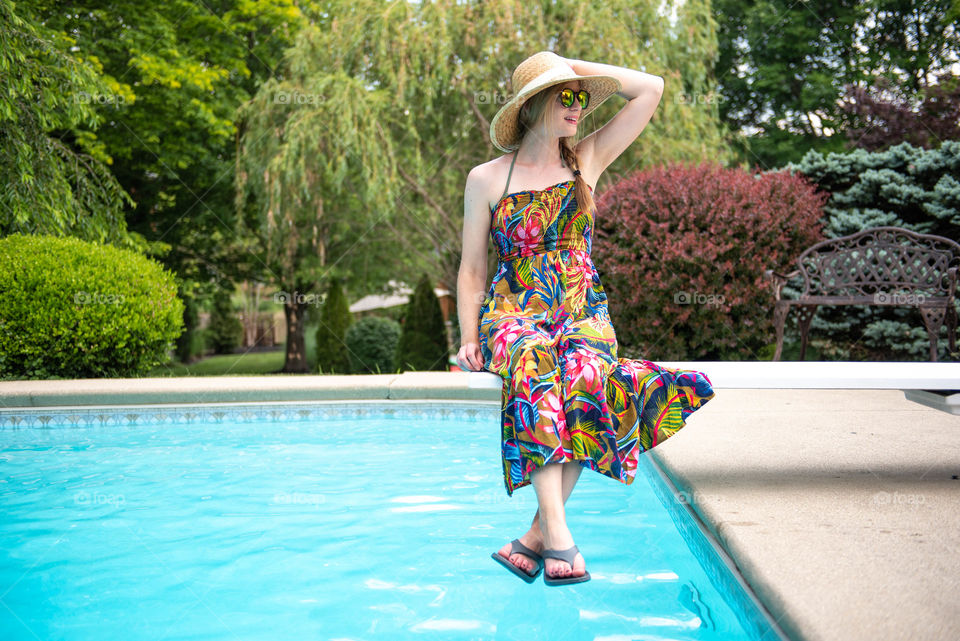 Young millennial woman wearing a dress and flip flop sandals while sitting on a diving board of a swimming pool in the summer