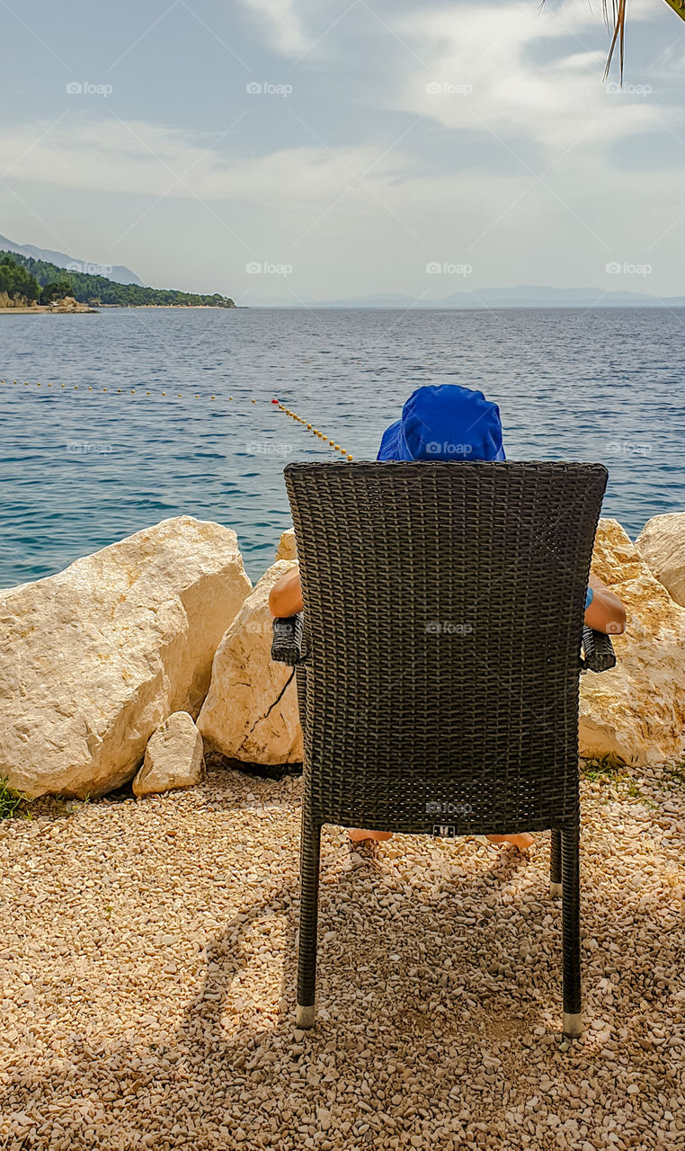 A boy sitting in a chair in the summer on the seashore and looking into the distance.  Summer holiday, relax