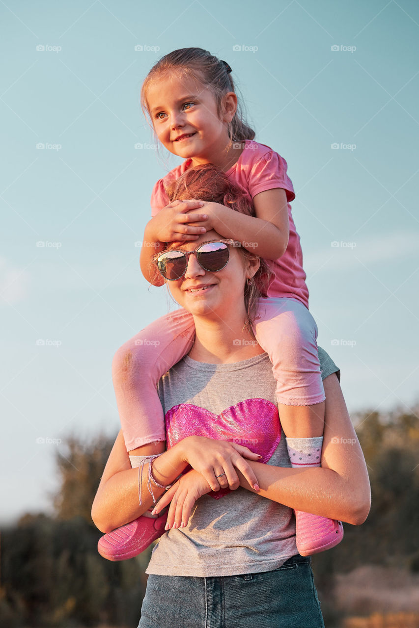 Sisters enjoying piggyback ride. Teenage girl carrying her younger sister up on the back and shoulders spending time playing together outdoors in the countryside. Candid people, real moments, authentic situations