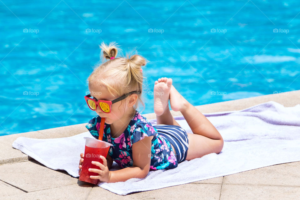 Cute little girl with blonde hair drinking juice near swimming pool 