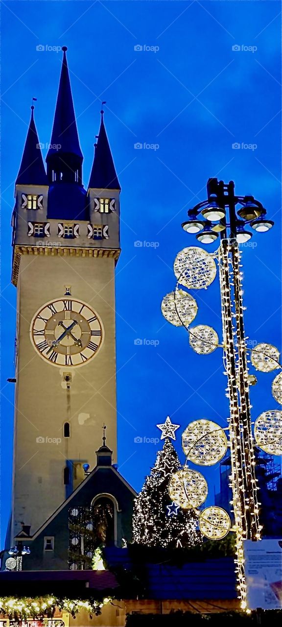 This is the medieval clock tower of “Straubing”, “Bavaria”next to illuminated Christmas decorations for the “Christkindlmarkt”, a popular Christmas fair on the “Theresienplatz”, one of two main squares in the inner city. 2023. Hypnotic Productions