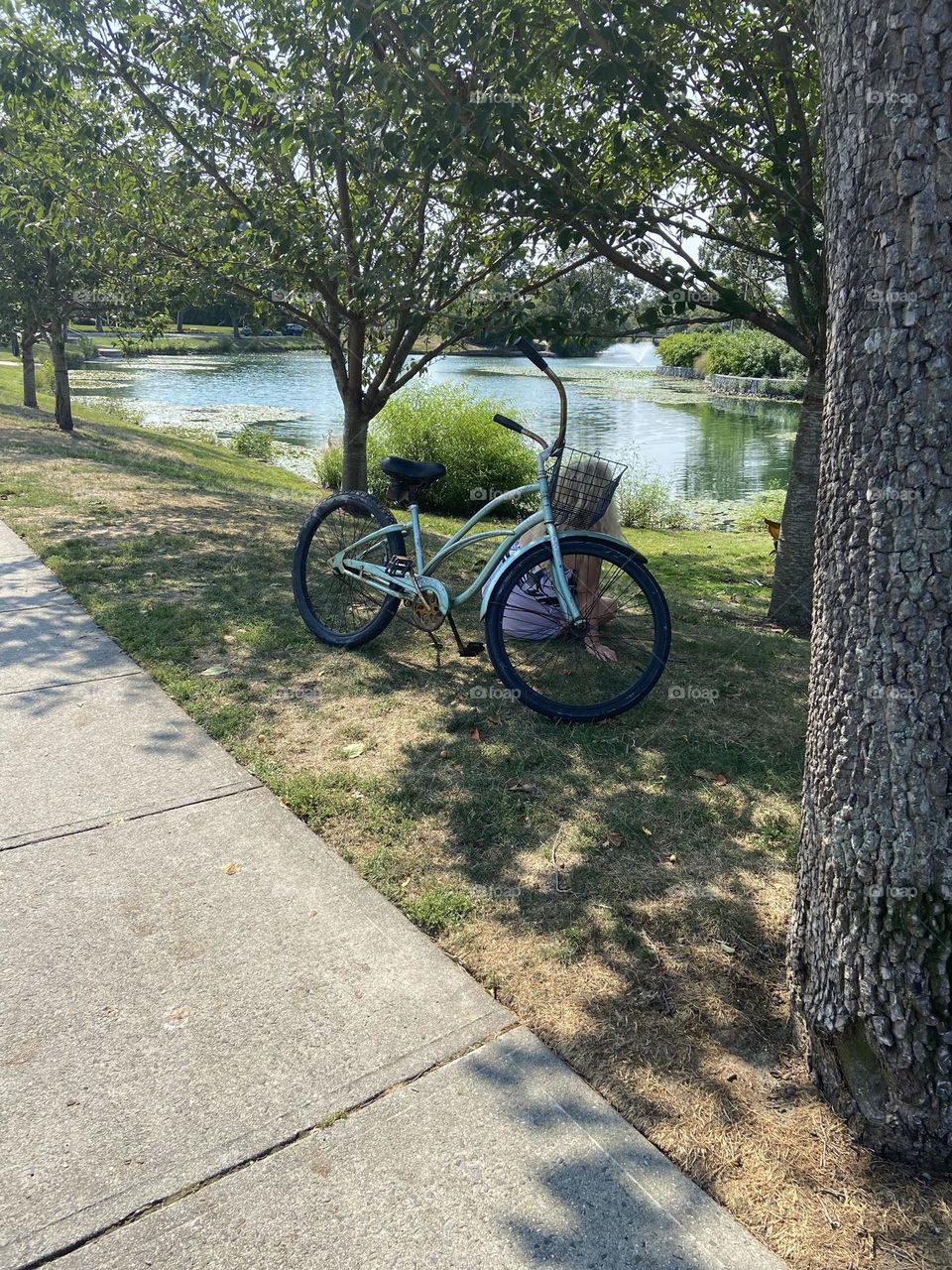 A woman sits on the grass in front of her bike at a local park. It’s a good spot to rest and view the lake before riding back home. Trees provide shade and shadows, and sunlight glimmers on the water. 