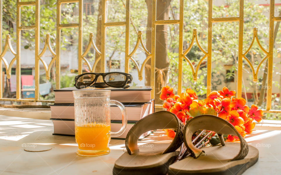 Modern women essential accessories for sunday weekend activities on wooden table. Sunglasses placed over Books, flip flops, a glass cocktail and flower bouquet with natural summer sunlight coming into