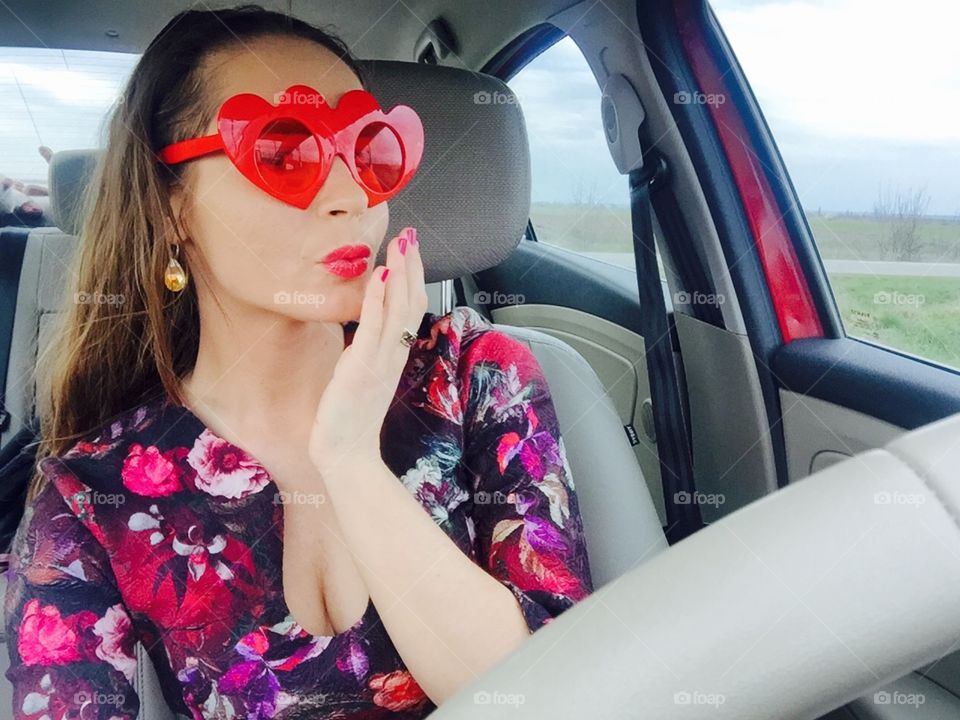 Selfie of woman in the car wearing heart-shaped sunglasses blowing a kiss