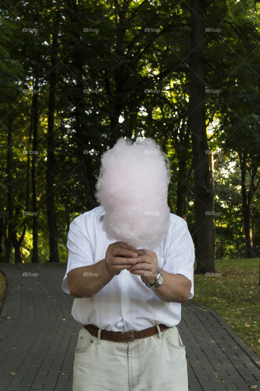 An elderly man of retirement age with wrinkles stretched out his hands and covered his face with pink cotton candy