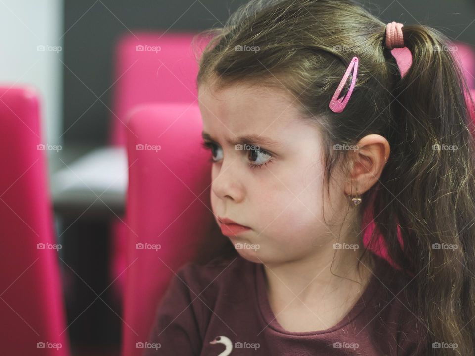 Portrait of one beautiful caucasian little brunette girl with ponytails on her head looks sadly and resentfully to the side, standing in a cafe in the afternoon, close-up side view. The concept of sad people and offended children.