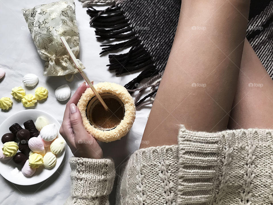 Young woman in cozy sweater drinking hot coffee and eating colorful candies in bed