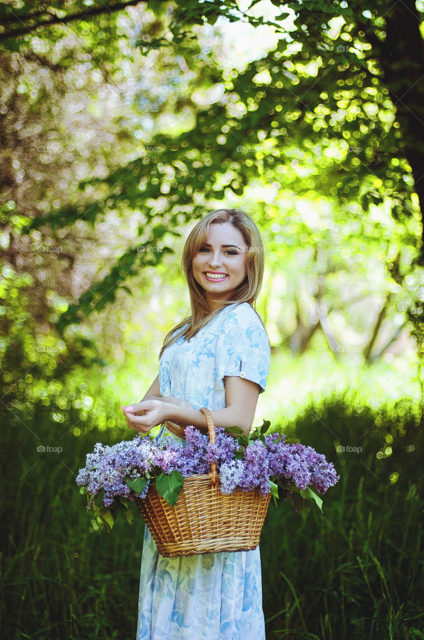 Beautiful young woman with basket of purple lilacs walking in green spring garden in may.