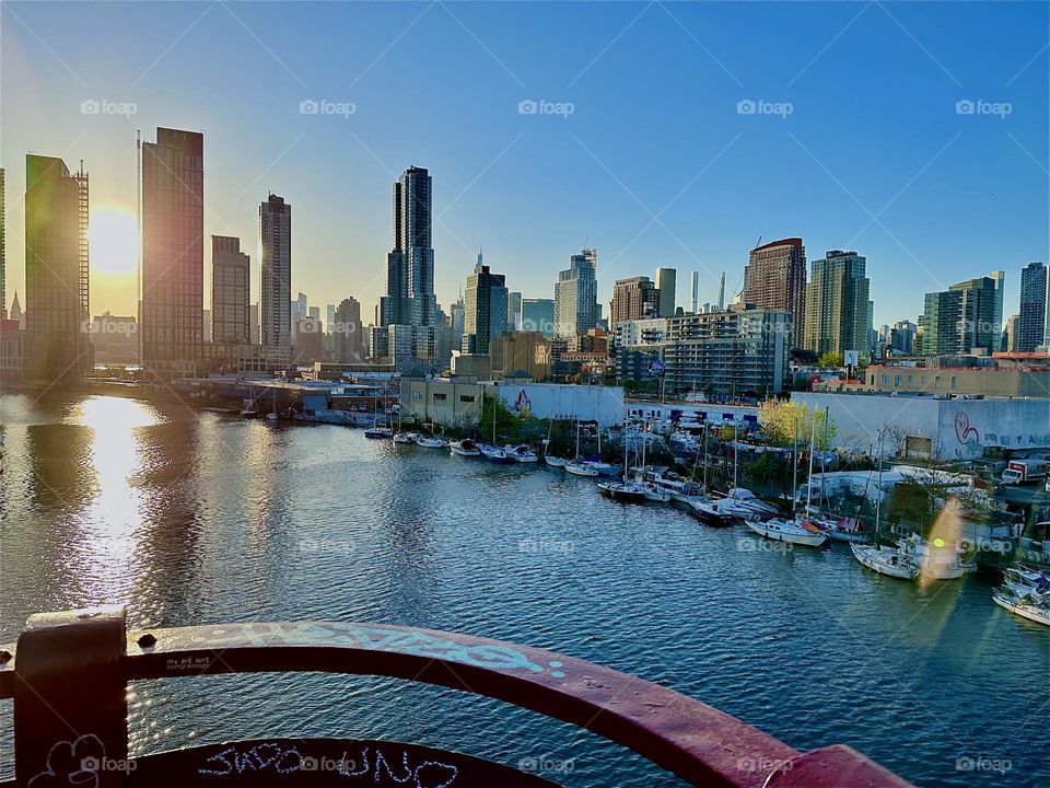 This is “Newtown Creek” seen from the “Pulaski Bridge” that connects “Greenpoint”, Brooklyn to LIC, Queens in the golden light of the setting evening sun with its many boats. In the distance we can see “Manhattan”. 2024. Hypnotic Productions