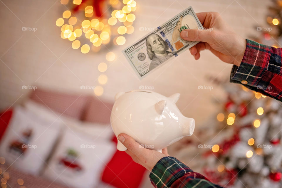 piggy bank with 100 dollars banknote in festive New Year atmosphere of scenery.  Magical bokeh with Christmas tree and bright lights.