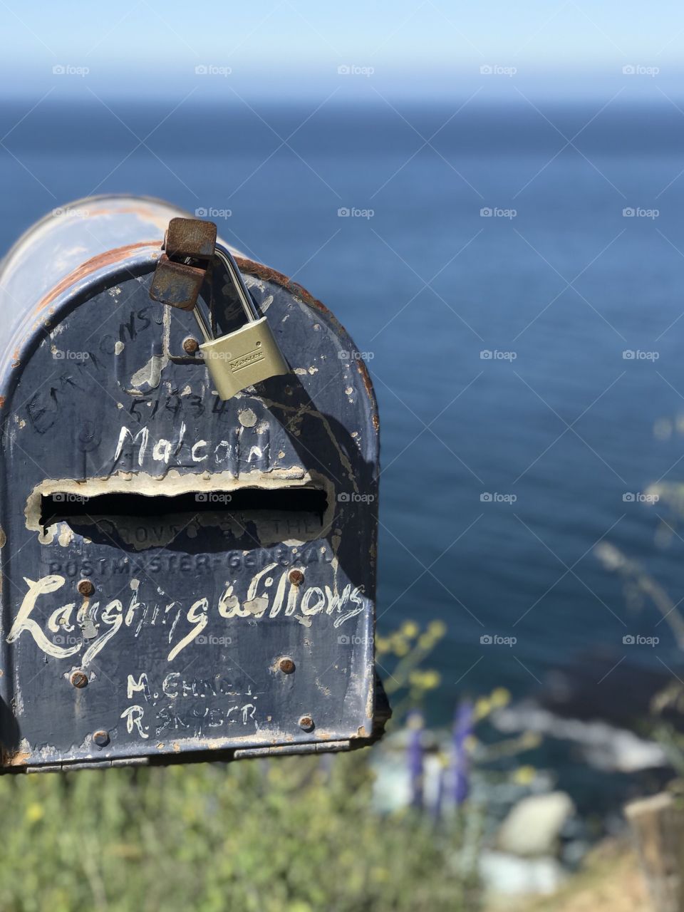 A detailed look at an ordinary mailbox. The aged and rusted metal and chipped paint create a work of art that is accentuated by the blue ocean behind it. 