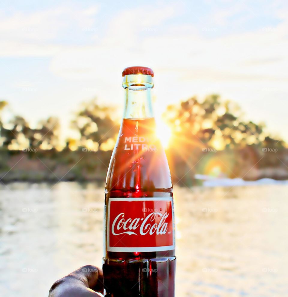 holding a bottle of soda pop as the sun goes down by River's Edge
