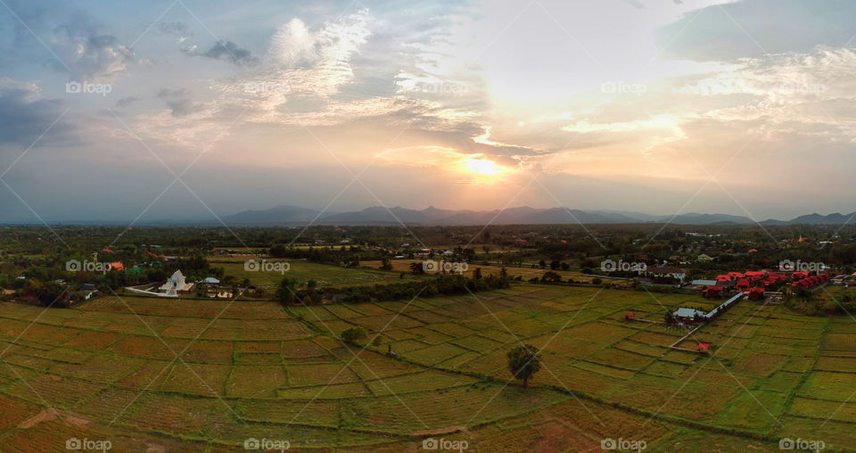 Beautiful Panoramic aerial view of village in the countryside farming area of Chiang Mai, Thailand with rice fields, houses, buddhist temple, forest and mountain during the evening sunset.