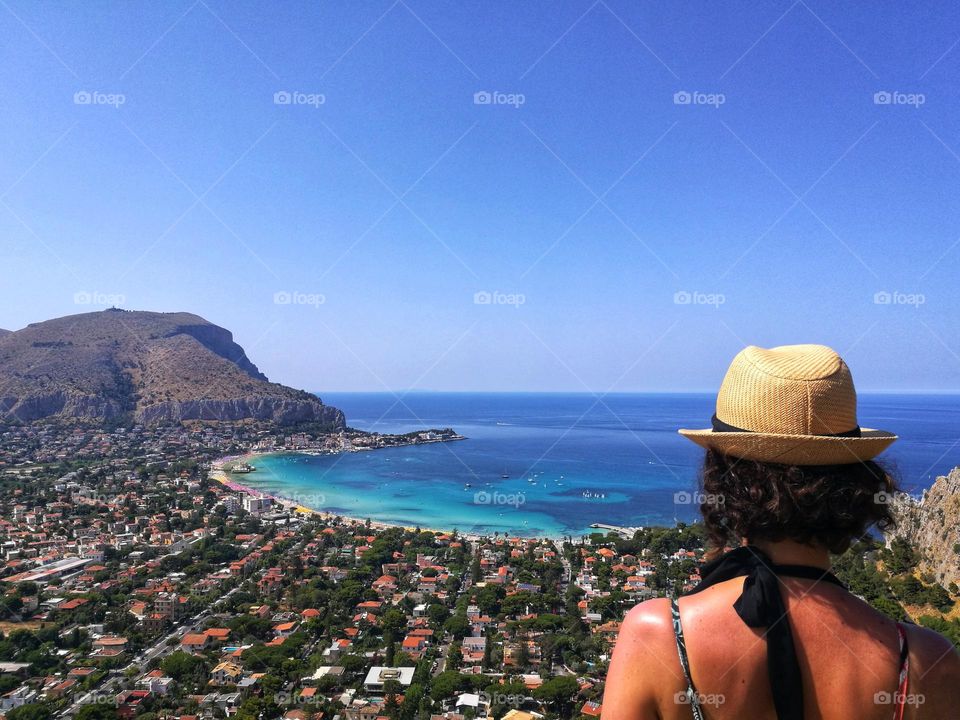 woman from behind with hat observes the blue sea of ​​Mondello, Sicily from above