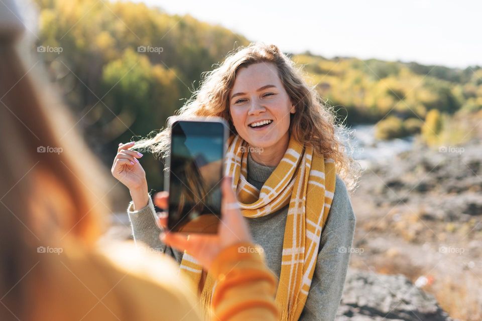 Young beautiful women travellers friends taking photos on mobile phone on view background of mountains and river, hiking on the autumn nature