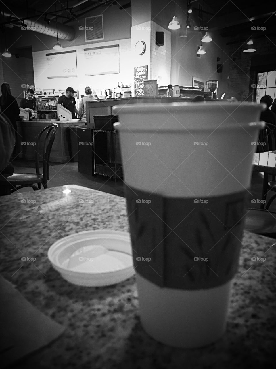 Hanging at the coffee shop.  There’s great people watching there.  Black and white photo with coffee cup on the foreground, blurred. 