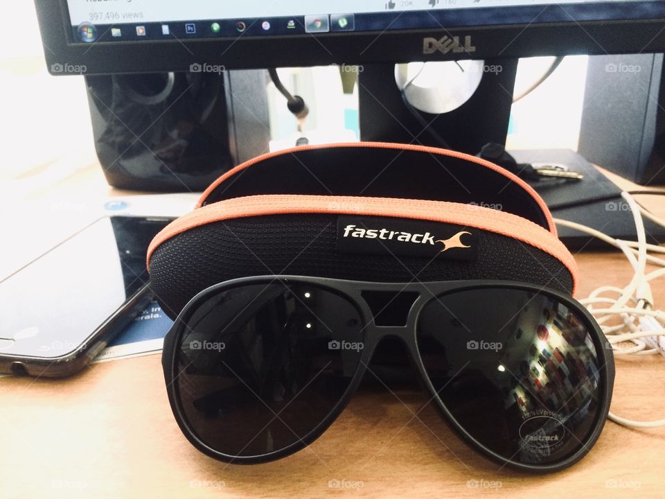 A fastrack sunglass on the table with its cover and ny earphone nearby on the computer table