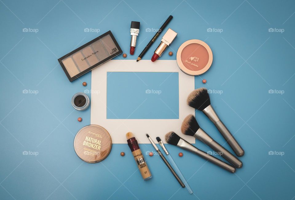 Cosmetics set of nude eyeshadow palette, round shape face powder box, red lipsticks, black soft eyebrow shadows, makeup brushes and eyeliners with white paper frame ledat in the center on a blue background with copy space in the center, flat lay clos