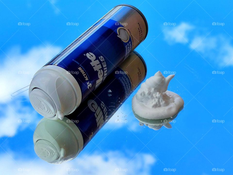 On the mirror surface lies a blue bottle with a white inscription: "Gillette series" and white fluffy foam.  Objects and a blue sky with white clouds are reflected in the mirror surface