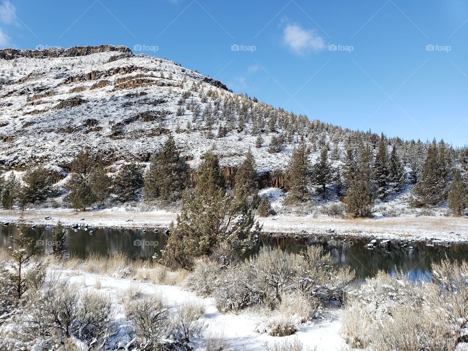 The snow covered banks of the Crooked River with juniper trees and a hill in the background on a winter day in Central Oregon with bright blue skies.