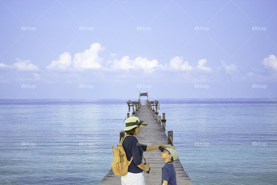 Mother shoulder backpack and son walking on wooden bridge pier boat in the sea and the bright sky at Koh Kood, Trat in Thailand.