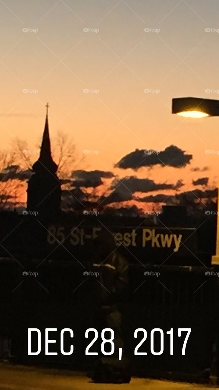Morning dawn at the train station in Queens New York.The sky is orange . A silhouette can be seen in front of the train stop sign. 