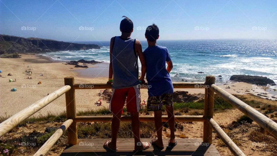 Two young boys standing on a wooden platform, admiring the beautiful beach scenery below them