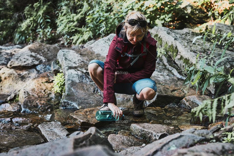 Woman taking pure water to bottle from mountain stream during trekking in mountains. Hiker crouching on rocks, filling bottle up with cold mountain water. Enjoying the outdoors in the summer trip vacation