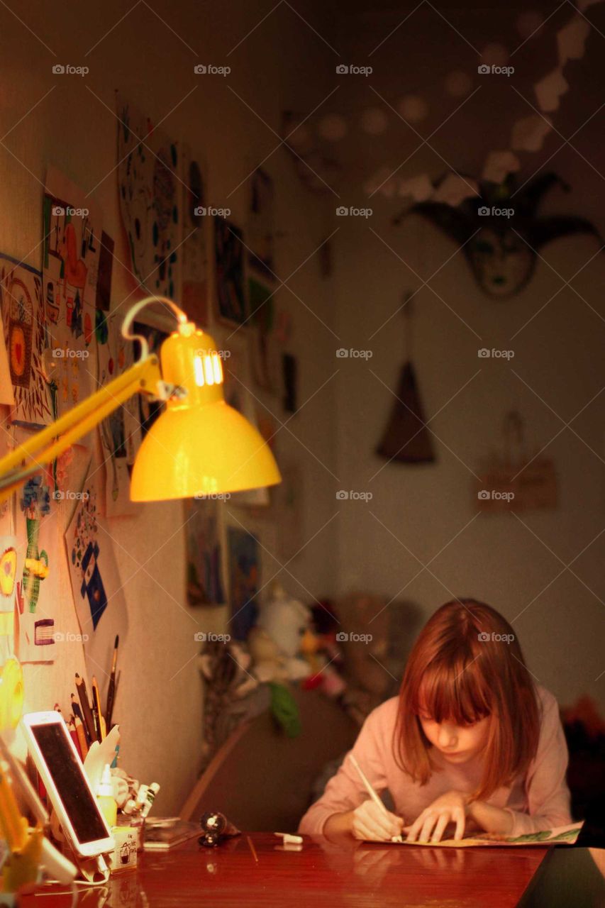 Little girl writes at the table under the light of a desk lamp