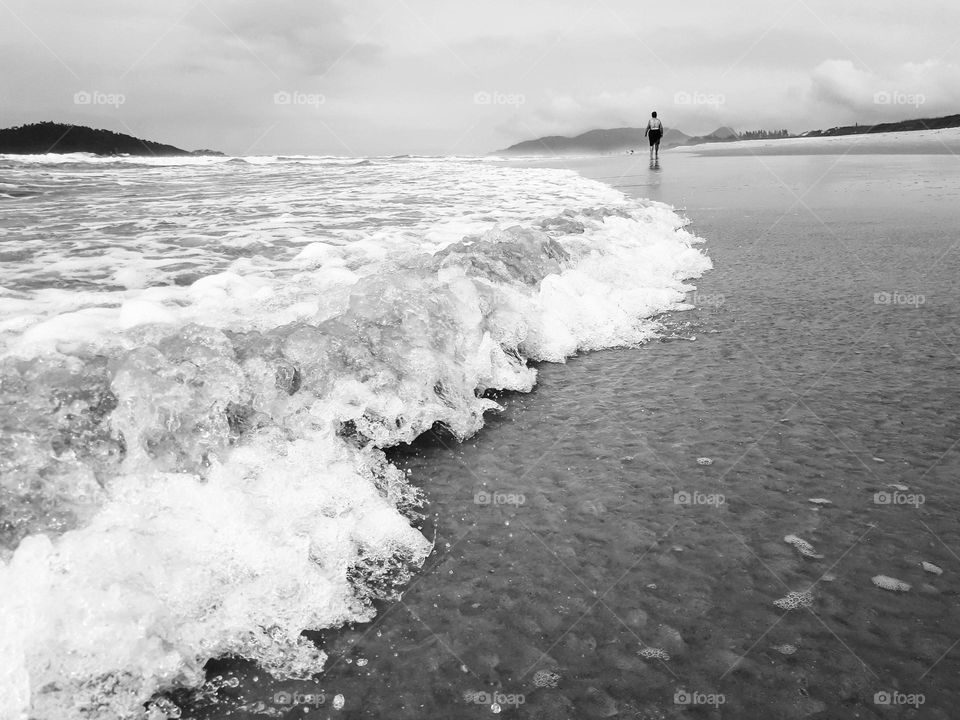 Image with angle and focus on the sea waves moving along the beach. In the background, a lady walks on the beach, observing the natural beauty around her.
