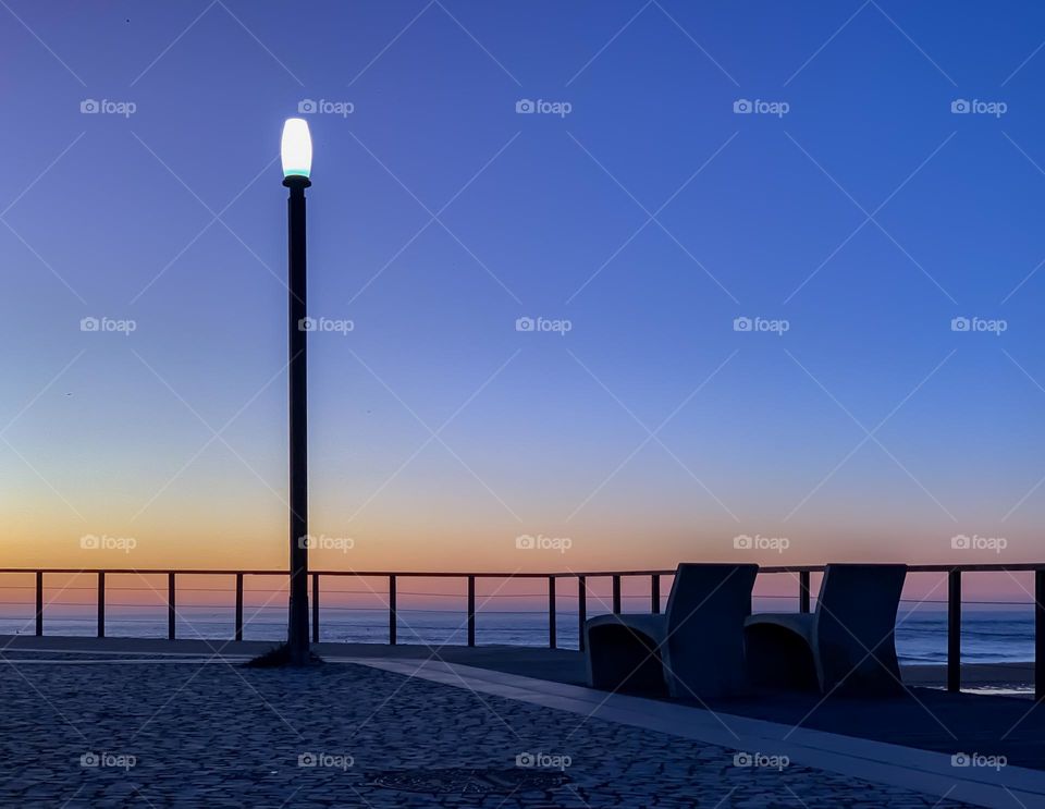 A twilight blue sky with two benches overlooking the sea next to a street light