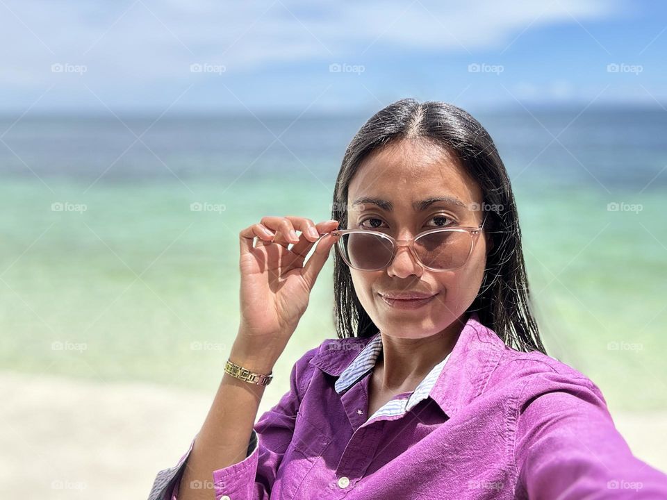 Portrait of a woman holding reflective eyeglasses wearing purple shirt with sky and ocean tropical beach background. 