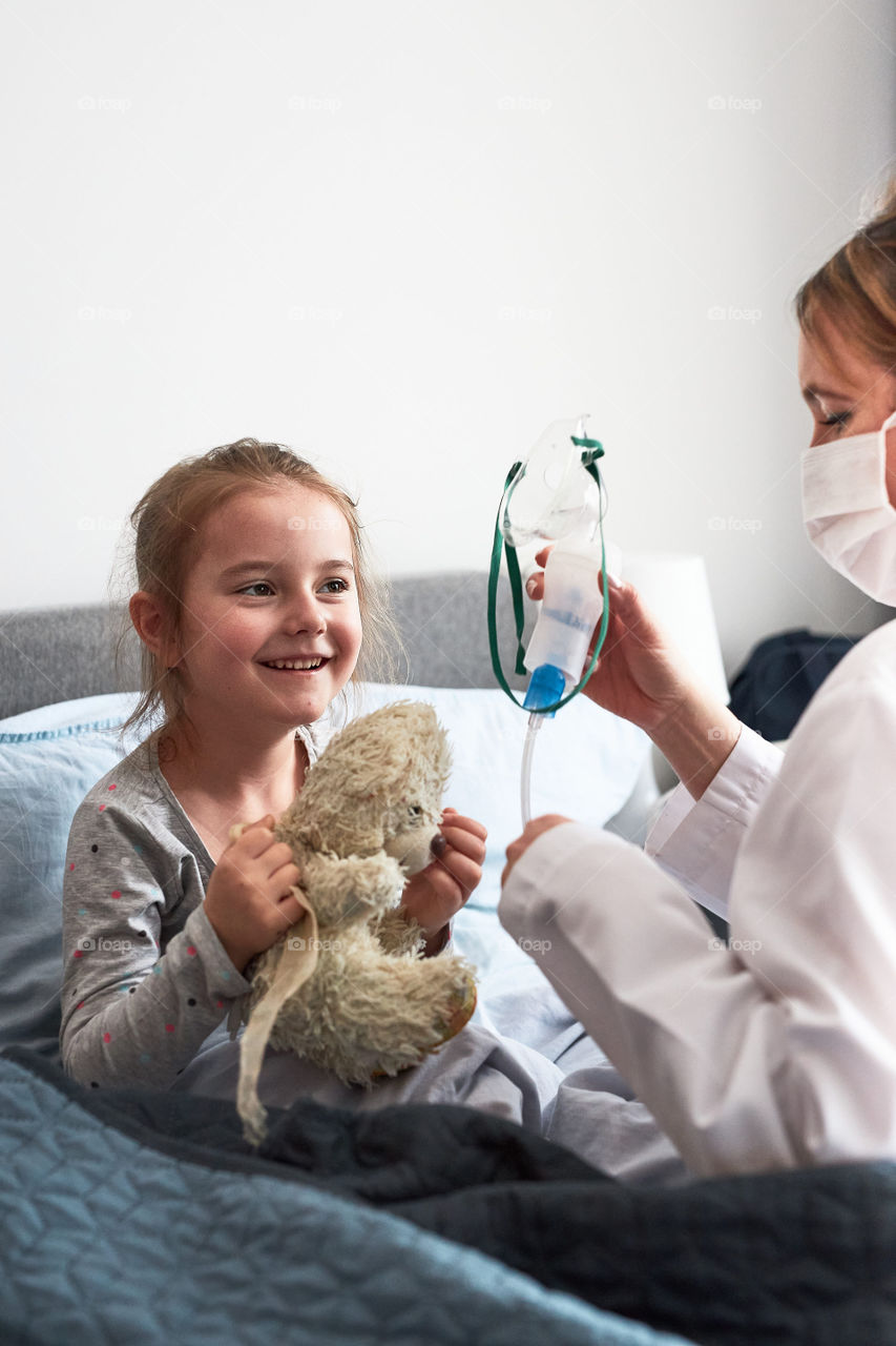 Doctor visiting little patient at home. Child having medical inhalation treatment with nebuliser. Woman wearing uniform and face mask