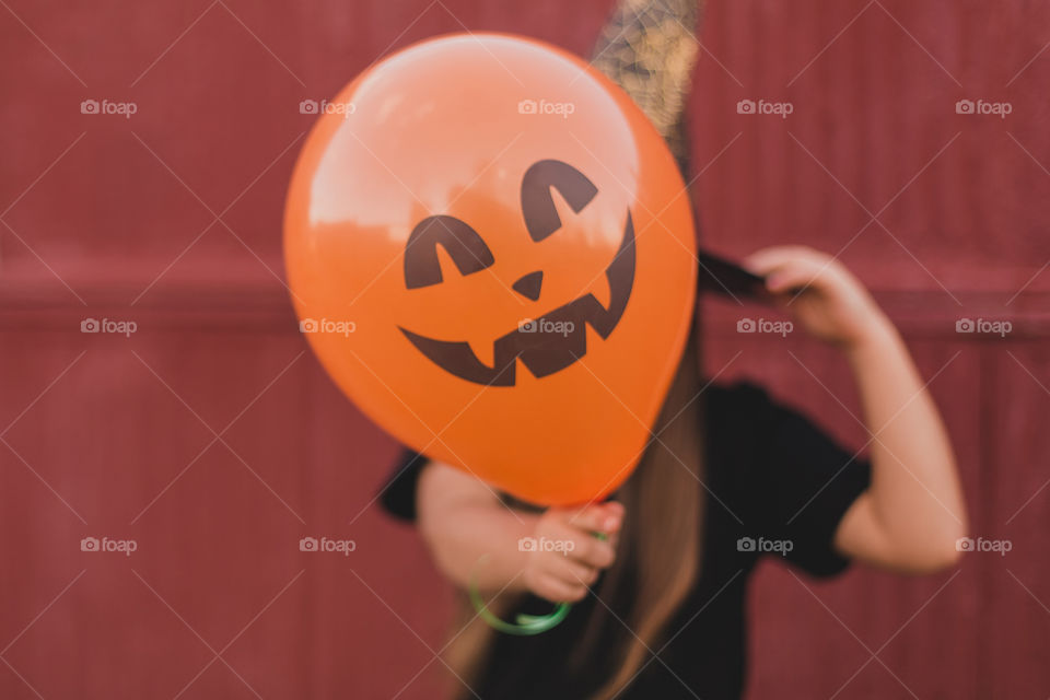 A little girl dressed as a witch for halloween holds a pumpkin-shaped balloon in her hands.