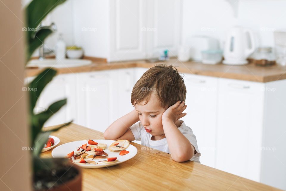 Cute toddler boy sitting with puncakes with berries in kitchen at the home. Child doesn't want to eat