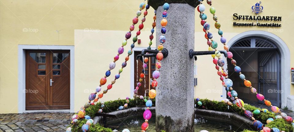 An antique fountain is being decorated for “Easter” with a round plant reef and multicolor eggs on strings in the beautiful city of “Regensburg” by the “Danube” just north of “Bavaria” in the “Oberpfalz”. 2023. Hypnotic Productions