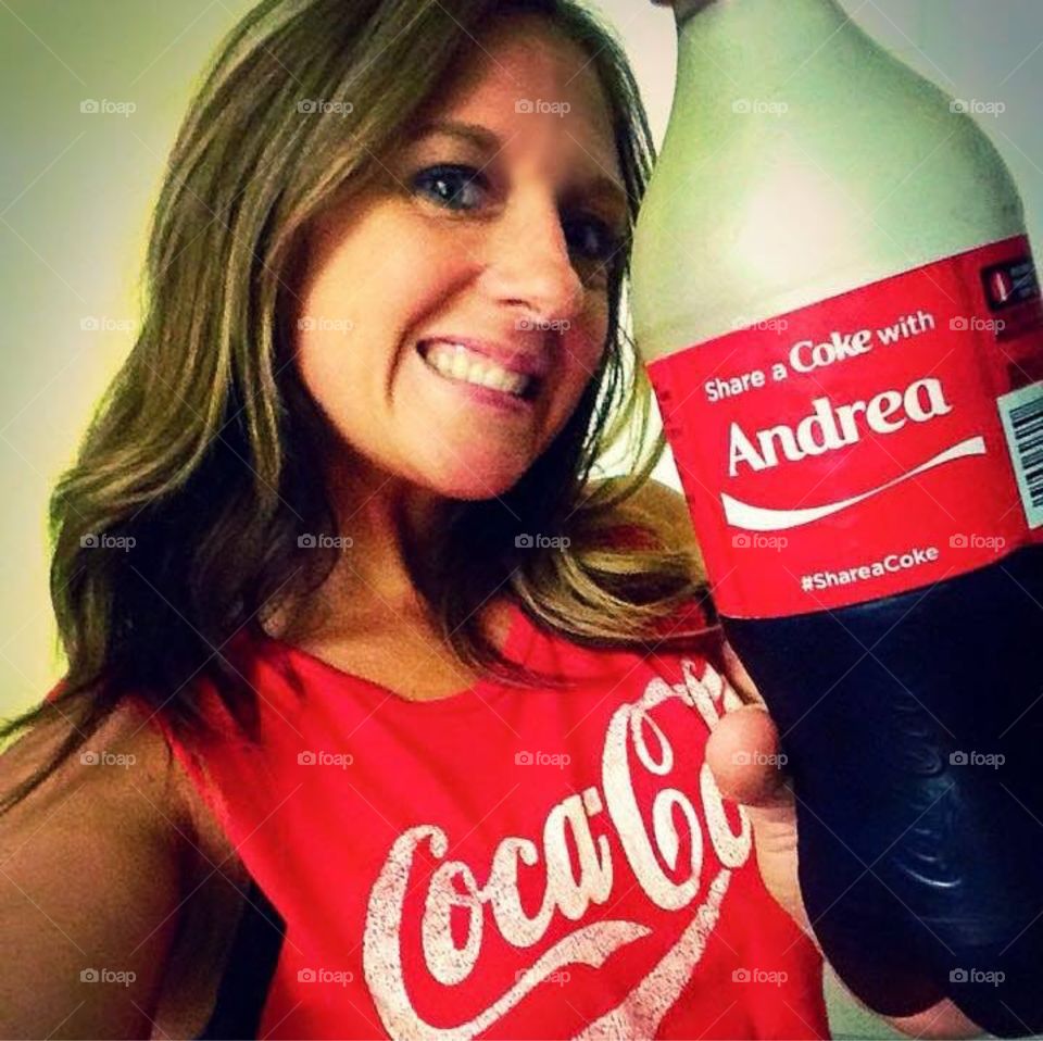 Share a Coke. Woman, wearing a Coke shirt, holding up a "Share a Coke" with her name on it. 