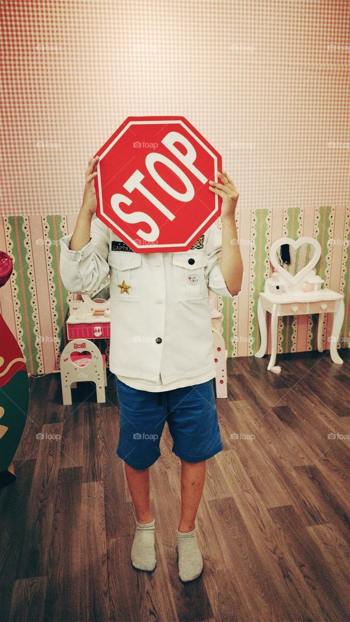 A person wearing a white shirt and blue shorts is holding a stop sign. This person is standing in a room with wooden floors and pink walls.