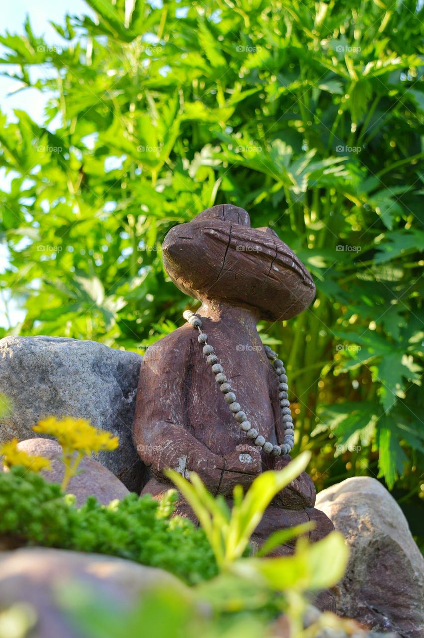 a wooden frog in a meditative pose with Buddhist monk beads around its neck. the figure is among the stones and grass against the background of green bushes