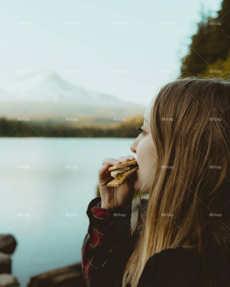 woman standing and eating food facing body of water viewing mountain.