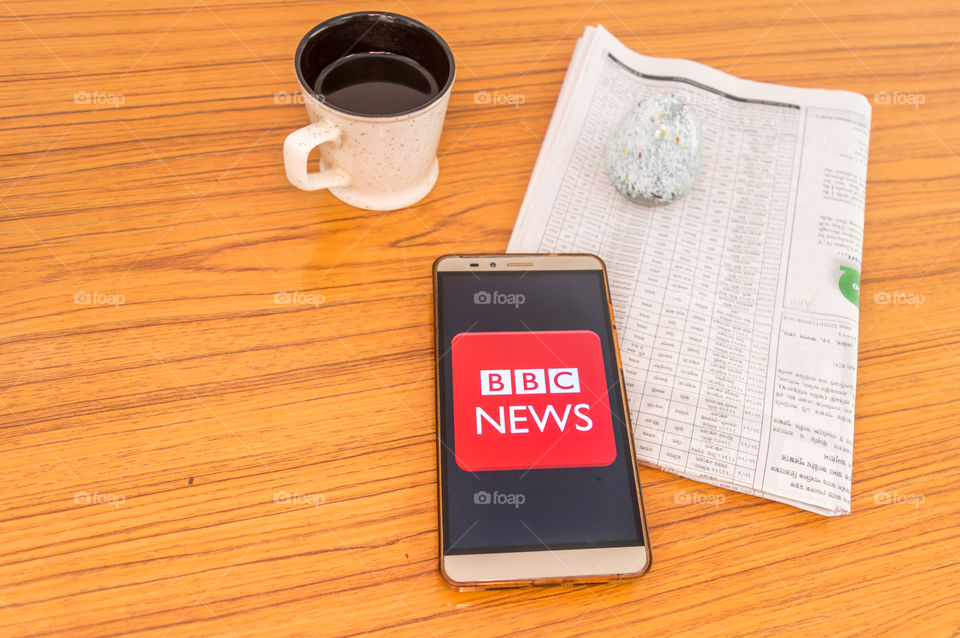 Kolkata, India, February 3, 2019: BBC news app (application) visible on mobile phone screen beautifully placed over a wooden table with a newspaper and a cup of coffee. A Technology Product Shoot.
