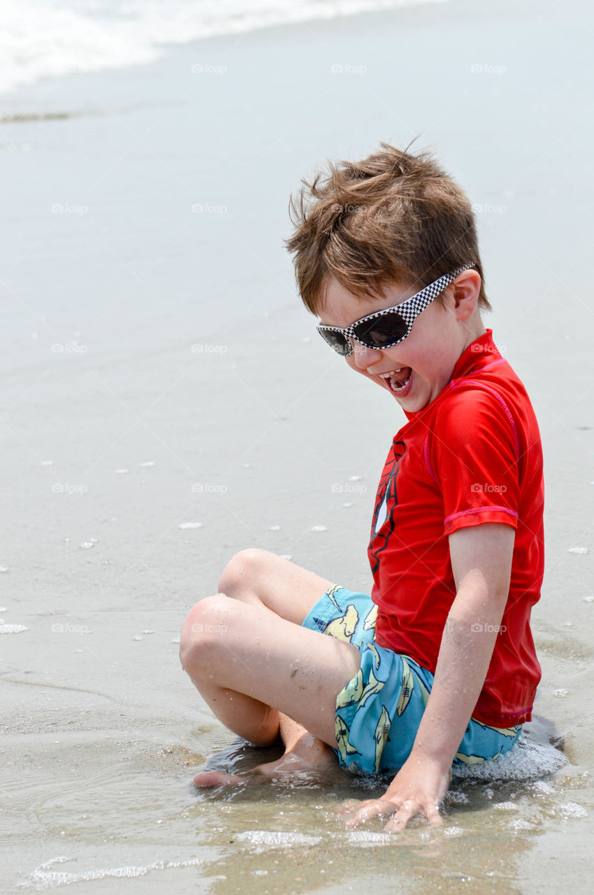 Young boy sitting at the shore of the beach and laughing with joy at the waves