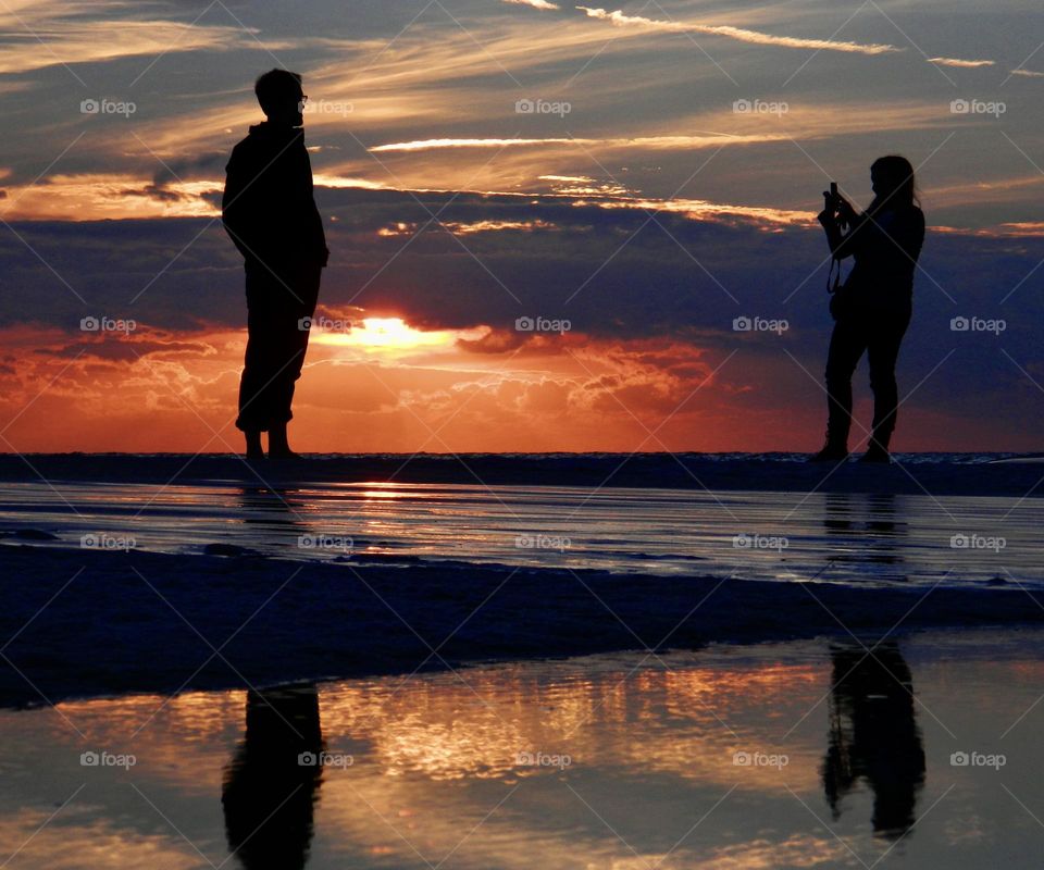 Battle: Summer vs Spring: A couple of vacationers having fun taking photos of each other during a colorful descending sunset on the Gulf of Mexico