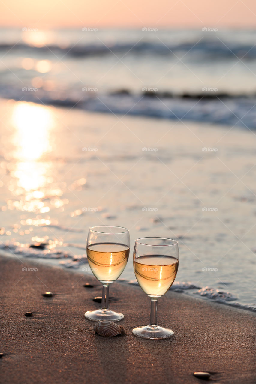 Two wine glasses with white wine standing on sand on beach. Sea waves in the background